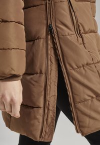 Brun polstret jakke med to lodrette lynlåse delvist åbne, som viser sorte bukser nedenunder, og en persons venstre hånd hviler ved siden af jakken.