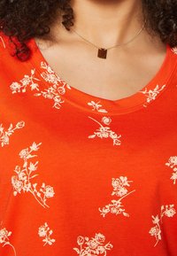 Orange stof t-shirt med rund hals, prydet med et hvidt blomstermønster, glat tekstur og et minimalistisk, rektangulært halskæde i guld.