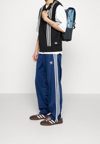 Suéter negro sin mangas con rayas blancas sobre una camisa blanca, pantalones deportivos azul marino con rayas blancas y zapatillas marrones con detalles blancos.