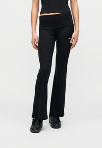 Μαύρο πλεκτό παντελόνι με καμπάνα και ψηλή μέση, με μικρό λευκό λογότυπο Nike. Φορεμένο με μαύρα αθλητικά παπούτσια, πάνω σε λευκό φόντο.