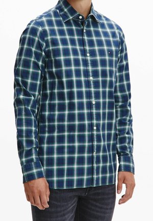 Homme portant une chemise à carreaux bleue et verte à manches longues avec un petit logo sur la poitrine et un jean foncé.