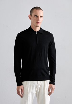 Black long-sleeve polo shirt with a zippered collar, made of soft material, paired with light-colored trousers, creating a contrast in colors.