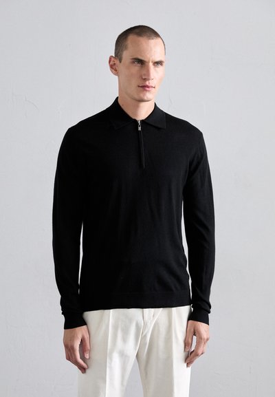 Black long-sleeve polo shirt with a zippered collar, made of soft material, paired with light-colored trousers, creating a contrast in colors.