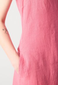 Ein pinkfarbene Leinenkleid mit einer taillierten Silhouette, seitlichen Taschen und einer strukturierten Oberfläche, die ein glattes, leicht zerknittertes Aussehen betont.