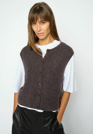 Woman with light brown hair wearing a dark gray buttoned vest over a white short-sleeve shirt and black leather pants, hands in pockets.