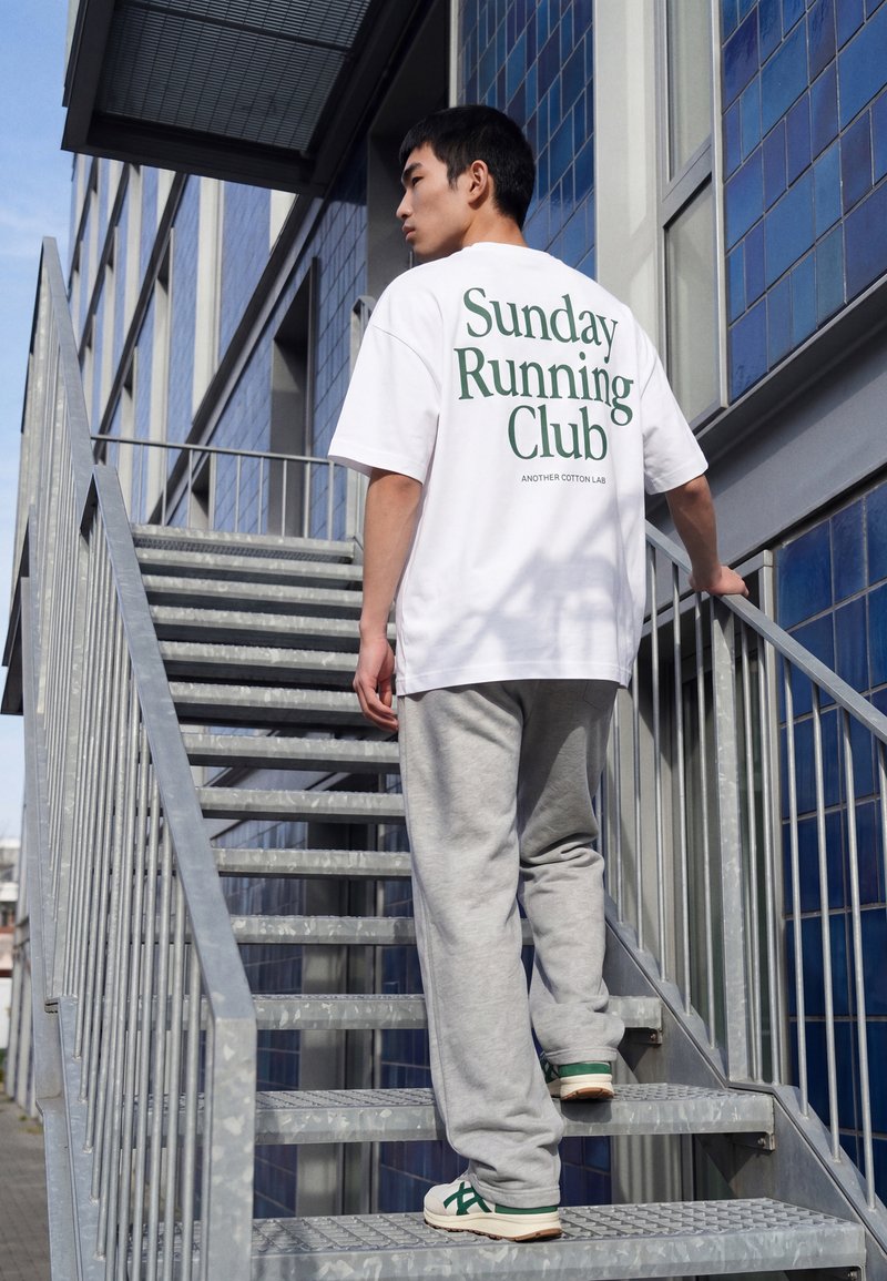 Giovane uomo che indossa una maglietta bianca "Sunday Running Club" e pantaloni della tuta grigi che sale una scala metallica esterna vicino a un edificio con piastrelle blu.