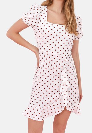 Woman wearing white dress with brown polka dots, puff sleeves, square neckline, and ruffle detail on the front hem.