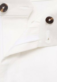Cuello de camisa de tela blanca con botones marrones y detalles de costura. Cuenta con una solapa con un ojal oculto. Textura suave y líneas limpias.