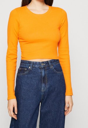 Femme portant un crop top orange vif à manches longues et un jean bleu taille haute, debout devant un fond clair uni.