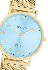 Montre ronde avec un cadran bleu clair, des accents dorés et un bracelet en maille dorée. Présente un design épuré et des index horaires minimalistes.