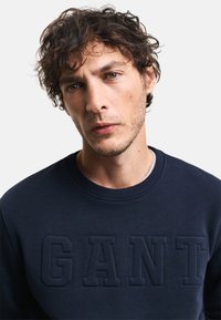 Sudadera azul marino con un cuello redondo acanalado y un logotipo "GANT" en relieve en la parte delantera. Fabricada con una tela suave con un acabado texturizado.