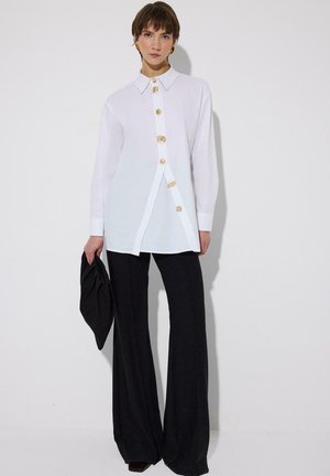 Ženska, ki nosi belo bluzo z gumbi različnih velikosti, črne široke hlače in drži črno torbico.