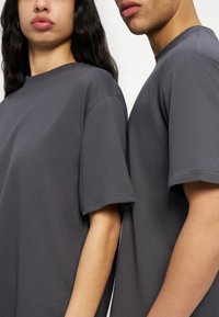 Δύο άτομα φορούν απλές σκούρες γκρι κοντομάνικες μπλούζες, στέκονται κοντά με ορατά τα χέρια και τους ώμους τους, εκφράζοντας ουδέτερες εκφράσεις.