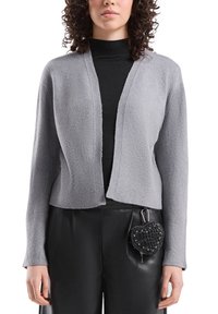 Szary krótki kardigan z dekoltem w szpic i prążkowanymi mankietami, noszony na czarnym topie. Do tego przypięty do spodni czarny dodatek w kształcie serca.
