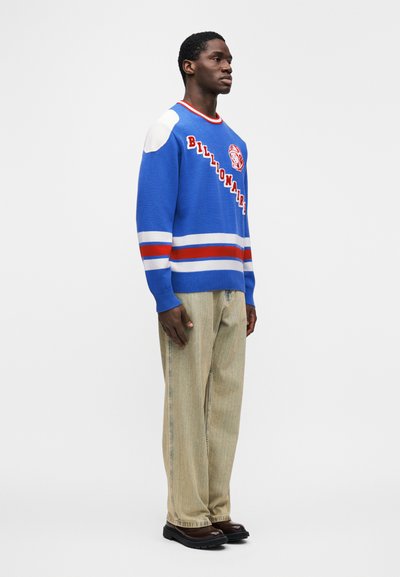 Μπλε πουλόβερ με κόκκινες και λευκές ρίγες, με το κείμενο «Billionaire» και λογότυπο· συνδυασμένο με μπεζ παντελόνια και μαύρα παπούτσια.