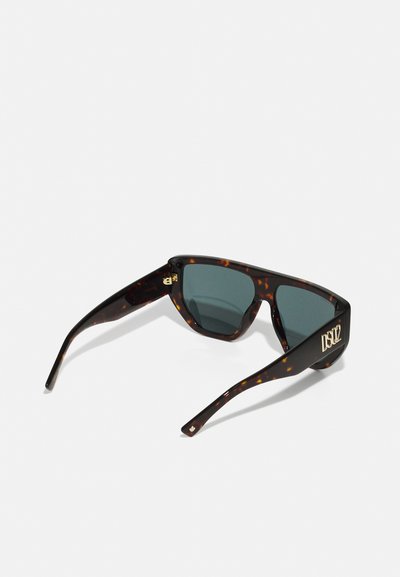 Óculos de sol tartaruga com armação geométrica, lentes escuras e um detalhe em logótipo dourado de lado. Textura suave, design elegante.