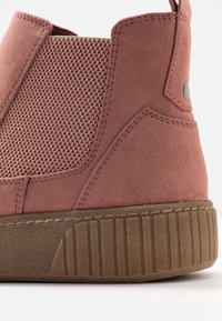 Primer plano de una bota ankle de ante rosa con panel lateral de elasticidad texturizada y suela gruesa estriada marrón.