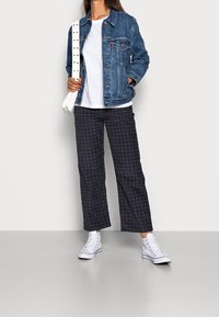 Farkkutakki vaalealla pesulla, valkoinen t-paita, ruudulliset tummansiniset housut ja valkoiset kengät punaisilla yksityiskohdilla. Malli poseeraa neutraalia taustaa vasten.