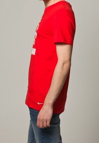 Czerwona koszulka z krótkim rękawem z bawełny, z graficznym nadrukiem z przodu i białym logo Nike na dolnym końcu. Noszona z niebieskimi dżinsami.