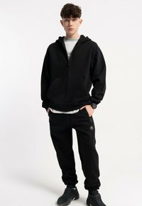 Czarna bluza z kapturem na zamek, w komplecie z dopasowanymi czarnymi spodniami dresowymi. Oba wykonane z miękkiego materiału; spodnie posiadają subtelny detal z logo.