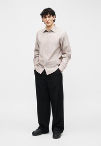 Mladý muž stojaci s rukami v vreckách, oblečený v béžovej košeli na gombíky a širokých čiernych nohaviciach na jednofarebnom pozadí.