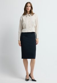 Beżowa krótka bluzka z guzikami, połączona z dopasowaną czarną spódnicą ołówkową. Modelka nosi czarne czółenka z ostrym noskiem. Gładkie tekstury w całej stylizacji.