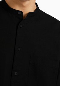 Camicia nera in lino con colletto alla mandarino, tasca frontale e bottoni neri, dalla texture liscia e dal design minimal.