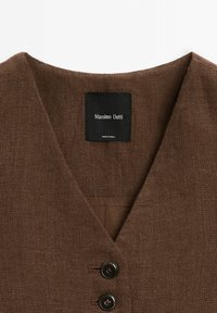 Brun linnesjuk med V-ringning, försedd med två svarta knappar och en svart etikett med texten "Massimo Dutti." Texturerat tyg med synlig väv.