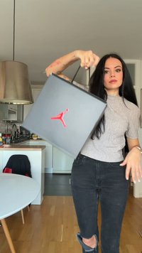 Ung kvinne med langt mørkt hår holder og svinger lett en svart Jordan-skoeske med en rød Jumpman-logo i et moderne kjøkken.