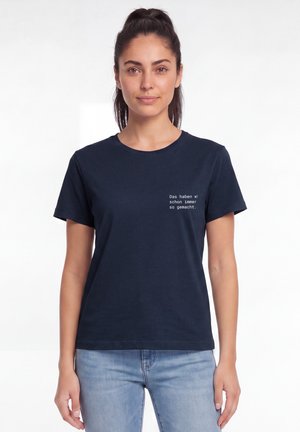 Vrouw met donker haar in een paardenstaart, die een marineblauw T-shirt draagt met witte Duitse tekst en lichtblauwe spijkerbroek, staand tegen een witte achtergrond.