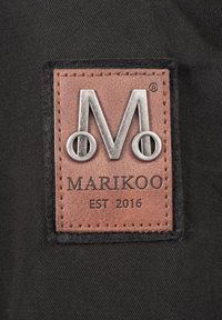 Étiquette en cuir marron avec un logo argenté "M" et le texte "MARIKOO EST 2016", cousue sur un fond en tissu noir.