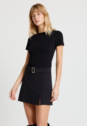 Donna con capelli biondi che indossa una maglietta nera aderente a maniche corte e una gonna nera con cintura e spacco frontale, in piedi davanti a uno sfondo semplice.