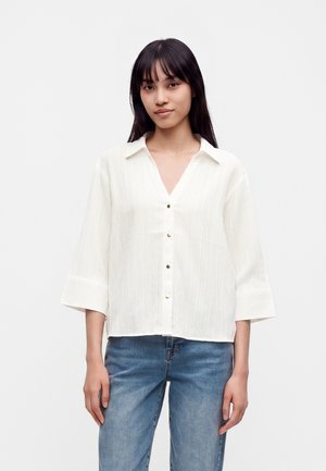 Femme aux longs cheveux foncés portant une chemise blanche boutonnée à manches trois-quarts et un jean bleu sur un fond uni.