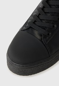 Schwarzer Sneaker mit einem strukturierten Lederobermaterial, glattem Gummizehenschutz, schwarzen Schnürsenkeln und einer strukturierten schwarzen Sohle. Nahaufnahme des Designs.