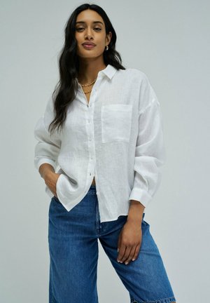 Mujer con el cabello largo y oscuro que lleva una camisa blanca holgada abotonada y vaqueros azules, de pie frente a un fondo liso gris claro.