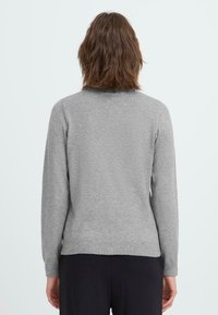 Frau mit schulterlangem braunem Haar, die einen schlichten grauen Langarmpullover und schwarze Hosen trägt, von hinten vor einem weißen Hintergrund gezeigt.