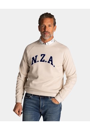 Man met grijze baard, gekleed in een beige sweatshirt met marineblauwe letters "N.Z.A.", een wit overhemd met patroon eronder, en een blauwe spijkerbroek, staand tegen een witte achtergrond.