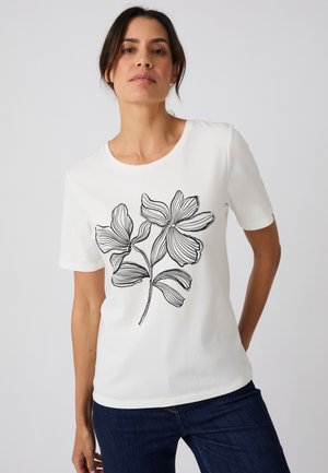 Femme portant un t-shirt blanc à manches courtes avec un motif floral dessiné au trait noir et un jean bleu foncé sur un fond clair uni.