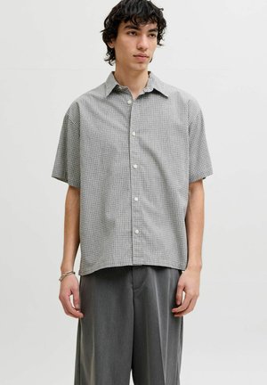 Jeune homme portant une chemise à manches courtes à carreaux grise à boutons et un pantalon ample gris foncé, debout devant un fond blanc uni.