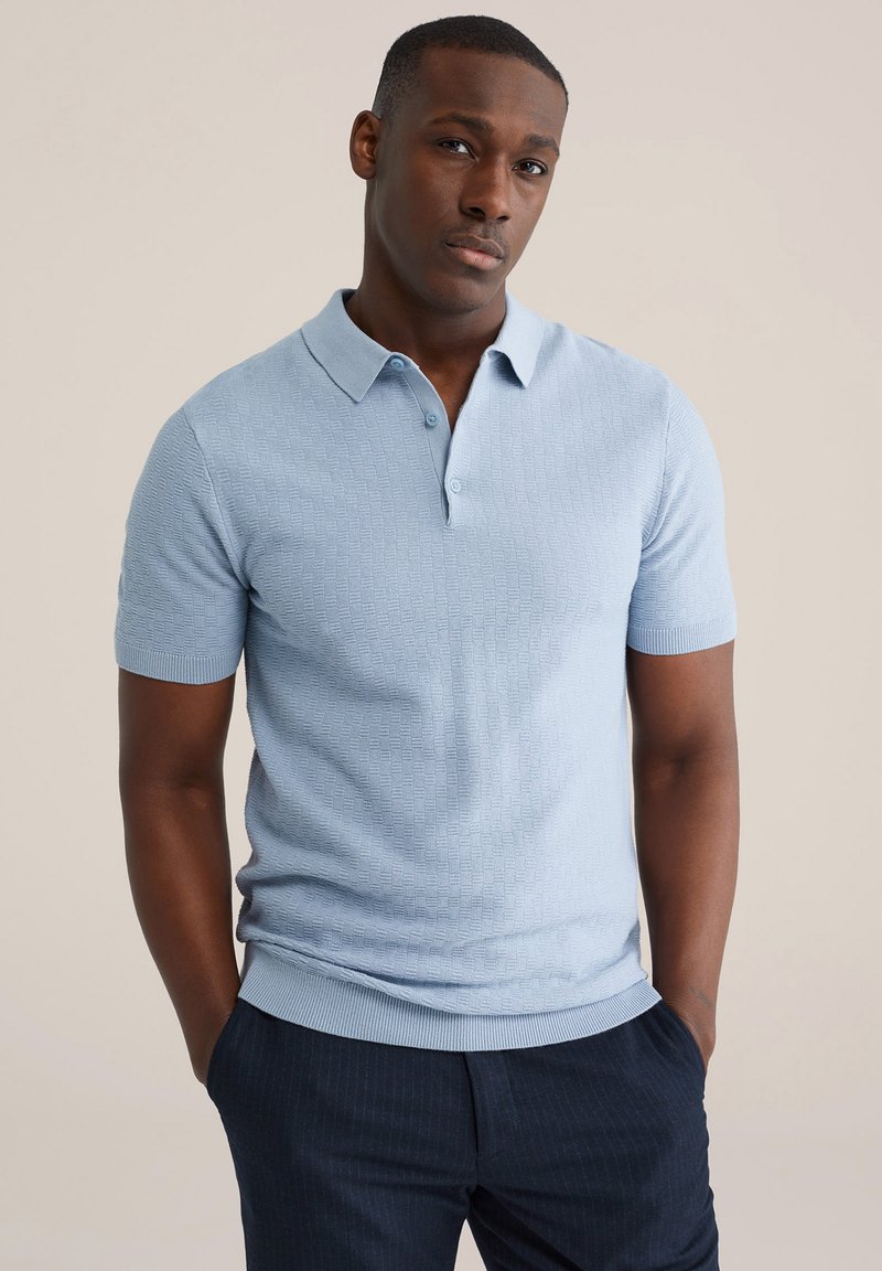 Polo a maniche corte azzurro chiaro con motivo a effetto tridimensionale, chiusura con bottoni e polsini a coste. Abbinata a pantaloni scuri a righe sottili.