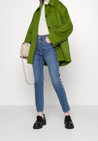 Abrigo de lana verde con botones grandes, blusa blanca, vaqueros de mezclilla azul claro y mocasines de plataforma negros; sosteniendo un bolso cruzado beige.