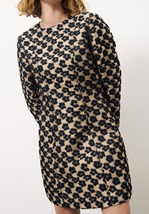 Kobieta w beżowej sukience z długim rękawem, ozdobionej czarnym, fakturowanym kwiatowym wzorem, stojąca na tle jednolitego białego tła.