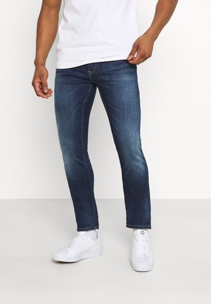 Homme portant un jean bleu foncé ajusté, des baskets blanches et un t-shirt blanc, debout devant un fond clair uni.