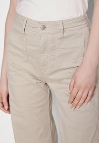 Pantalons taille haute beige avec deux poches plaquées à l'avant et un bouton en métal, portés avec une chemise blanche rentrée, montrant des mains reposant sur les côtés.