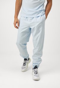 Gaiši zili sporta bikses no mīksta auduma ar elastīgu jostasvietu un piedurknēm, apvienotas ar melniem un baltiem sporta apaviem.