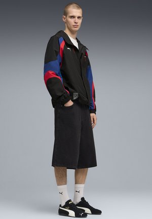 Junger Mann steht mit den Händen in den Taschen und trägt eine schwarze, rote, blaue Puma-Jacke, schwarze Shorts, weiße Puma-Socken und schwarze Puma-Sneaker.