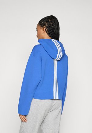 Blaues Hoodie aus weichem Material, mit einem gespaltenen Rücken-Design und drei weißen Streifen in der Mitte. Das Model trägt graue Hosen.