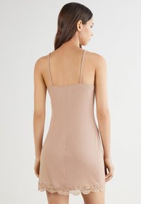 Vestido deslizante sin mangas en tono nude con tirantes delgados, que cuenta con un dobladillo de encaje en la parte inferior. Material suave, diseño ajustado con una textura lisa.