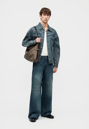 Jeune homme portant une veste en denim bleu et un jean à jambes larges, tenant un grand sac gris à bandoulière sur un fond blanc.