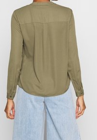 Oliwkowa koszula z długim rękawem, okrągłym dekoltem, subtelnym wycięciem z tyłu oraz mankietami zapinanymi na guziki. Gładka faktura i luźny krój.